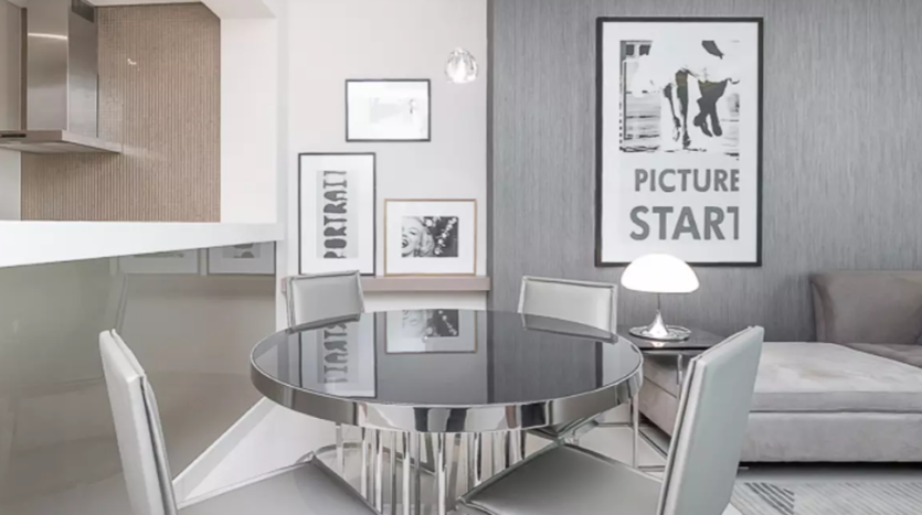 Coin repas moderne dans les tours Damac avec une table en verre, quatre chaises et des œuvres d&#039;art encadrées sur des murs gris. L&#039;espace s&#039;ouvre sur une cuisine et comprend une décoration minimaliste aux tons sourds.