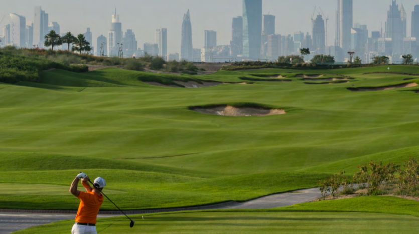 Un golfeur en chemise orange balance un club sur les greens luxuriants et vallonnés du parcours de golf de Dubai Hills avec l&#039;horizon d&#039;une ville, y compris d&#039;imposants gratte-ciel, en arrière-plan.