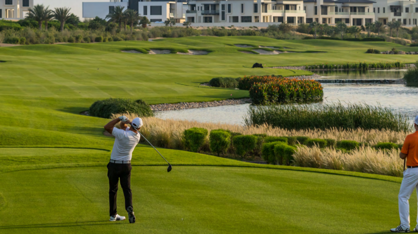 Un golfeur en chemise blanche balance un club sur le parcours de golf verdoyant de Dubai Hills avec un compagnon en observation orange, avec un fond pittoresque avec de l&#039;eau et un aménagement paysager.