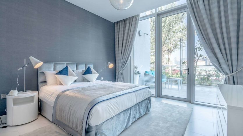 Une chambre moderne avec un grand lit, une literie bleue et grise, des murs et des rideaux gris. comprend des tables de nuit avec des lampes et une vue sur un balcon avec de la verdure à l&#039;extérieur.