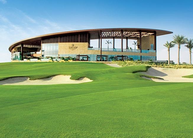 Club-house de golf moderne avec de grandes fenêtres en verre et un toit incurvé à Damac Hills, surplombant un parcours de golf bien entretenu avec des bunkers de sable et de l&#039;herbe verte.