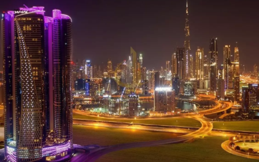 Vue nocturne d'un paysage urbain bien éclairé avec des gratte-ciel modernes, dont une tour avec un éclairage violet, et des routes courbes et éclairées. La Burj Khalifa et les Damac Towers Paramount sont visibles