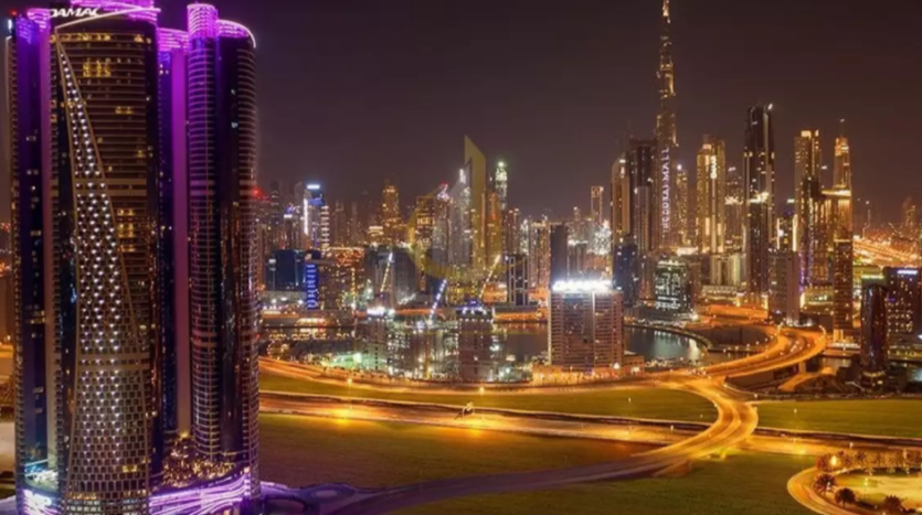 Vue nocturne d&#039;un paysage urbain bien éclairé avec des gratte-ciel modernes, dont une tour avec un éclairage violet, et des routes courbes et éclairées. La Burj Khalifa et les Damac Towers Paramount sont visibles