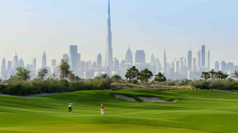 Deux golfeurs sur un parcours verdoyant avec un bunker au premier plan et l&#039;horizon des collines de Dubaï, avec le Burj Khalifa, en arrière-plan sous un ciel clair.