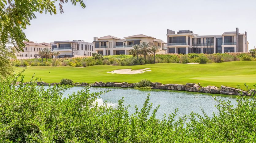 Un parcours de golf luxuriant avec un étang pittoresque au premier plan, entouré d&#039;une verdure vibrante et présentant une rangée de maisons modernes et luxueuses en arrière-plan sous un ciel dégagé sur les collines de Dubaï.