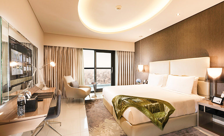 Une chambre d&#039;hôtel moderne dans les tours Damac comprenant un grand lit avec un jeté vert, un plafonnier rond, un coin bureau, un fauteuil et une vue sur la ville à travers la fenêtre. Les tons neutres et le design contemporain rehaussent