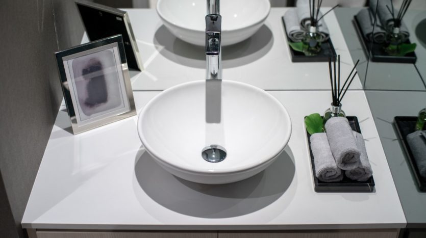 Un meuble-lavabo moderne avec un lavabo circulaire blanc, un robinet chromé et un miroir au mur. Les objets décoratifs comprennent un petit cadre de Wilton Park, une feuille verte et des serviettes grises soigneusement roulées