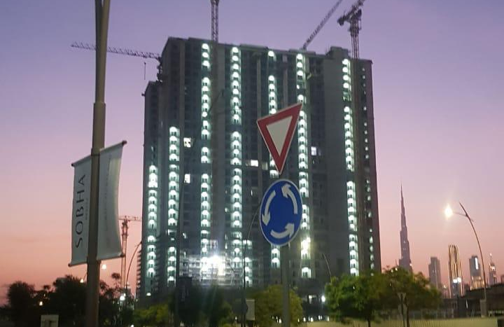 Paysage urbain au crépuscule observant de grands bâtiments en construction avec des grues à Creek Vistas Dubaï, le premier plan montre des panneaux routiers et une partie d&#039;une voiture, avec un horizon urbain et un lampadaire en arrière-plan.