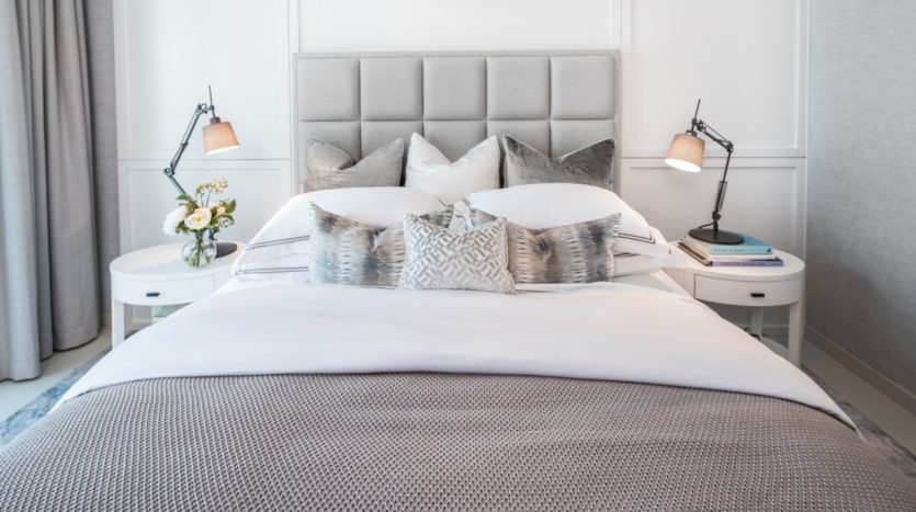 Une chambre bien aménagée à Wilton Park avec un grand lit doté d&#039;une tête de lit capitonnée grise, une literie blanche et grise, deux lampes de table élégantes de chaque côté et un vase de fleurs dessus.