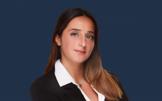 Portrait professionnel d&#039;une femme aux longs cheveux bruns portant un blazer noir sur une chemise blanche, idéal pour une agence immobilière de Dubaï, sur fond bleu.