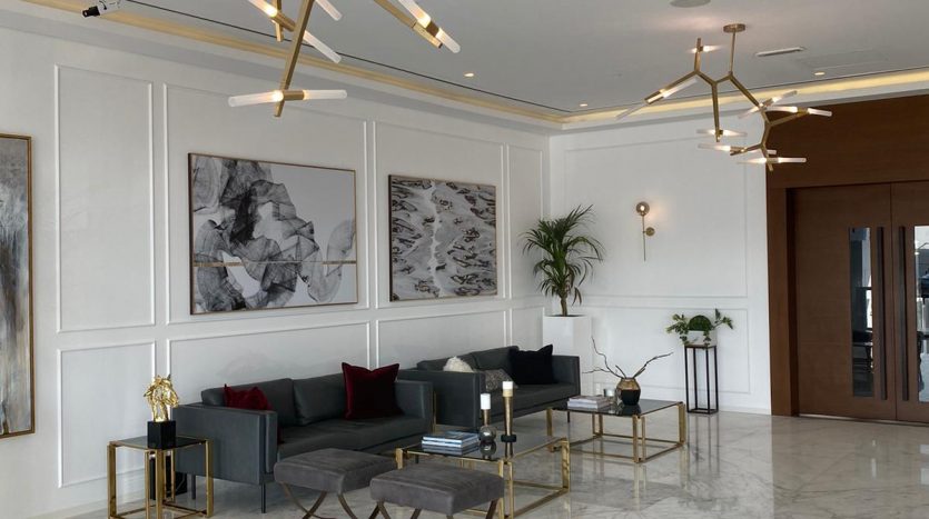 Lobby moderne et élégant avec sol en marbre blanc, grandes œuvres d&#039;art abstraites sur murs lambrissés blancs, sièges élégants gris et noirs, tables basses géométriques dorées et lustres dorés distinctifs offrant une vue imprenable sur Dubaï.