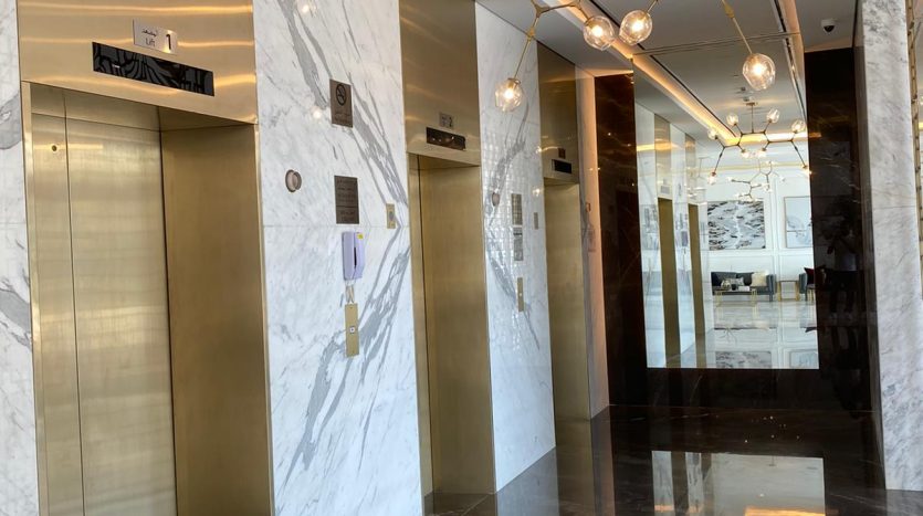 Hall d&#039;entrée moderne avec murs et sols en marbre poli, quatre ascenseurs avec portes métalliques et plafond décoratif avec luminaires sphériques, offrant une vue imprenable sur Dubaï.