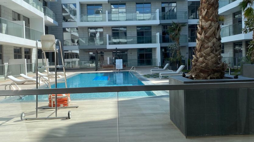 Un complexe d&#039;appartements moderne à Dubaï avec une piscine bleu clair au centre, entourée de chaises longues et d&#039;un grand palmier. Les bâtiments ont une façade blanche contemporaine avec des balcons offrant des vues privilégiées.