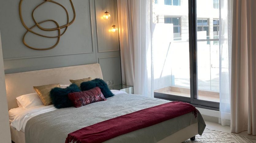 Une chambre moderne au design minimaliste avec un lit gris, des draps blancs, des coussins colorés, des murs bleu clair et une œuvre d&#039;art murale unique. La pièce dispose de grandes fenêtres offrant une vue imprenable sur