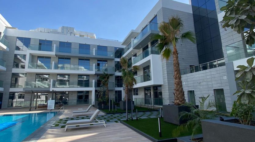 Bâtiment résidentiel moderne à Dubaï avec une piscine extérieure entourée de palmiers et de chaises longues, offrant une vue imprenable, un ciel bleu clair et une architecture géométrique.