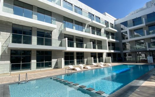 Un complexe d&#039;appartements moderne à Dubaï avec une grande piscine dans la cour, entouré de bâtiments blancs à plusieurs étages sous un ciel bleu clair.