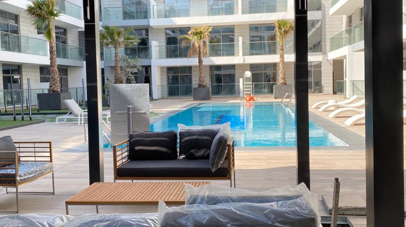 Un patio moderne avec une vue imprenable sur une piscine entourée d&#039;un immeuble résidentiel. Les caractéristiques comprennent des chaises en bois avec des coussins, un canapé et des palmiers luxuriants en arrière-plan.