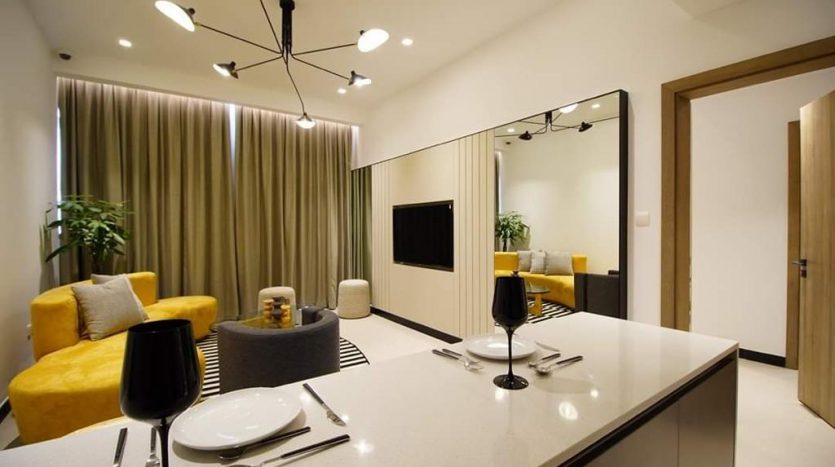 Salon moderne de l&#039;Aria Résidence Dubai avec un canapé jaune, une table à manger en verre, un lustre unique et un mur en miroir reflétant une télévision et une autre partie de la pièce.