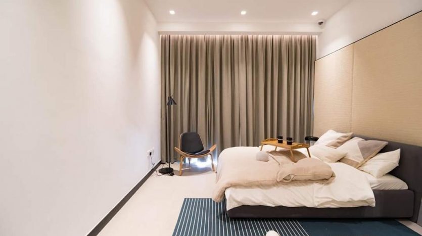 Chambre minimaliste moderne avec un grand lit recouvert de literie blanche, une petite chaise noire dans le coin et des rideaux beiges. La chambre présente un éclairage doux et un design élégant et épuré à la résidence Aria