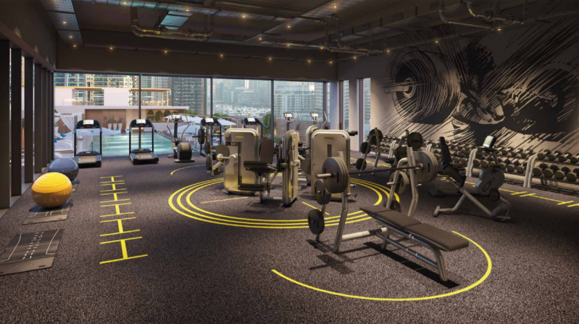 Intérieur de salle de sport moderne avec divers équipements d&#039;exercice, notamment des tapis roulants, des poids et des appareils de résistance. Le sol est marqué de lignes jaunes et les grandes fenêtres du Studio One Dubai Marina montrent un horizon urbain à l&#039;extérieur.