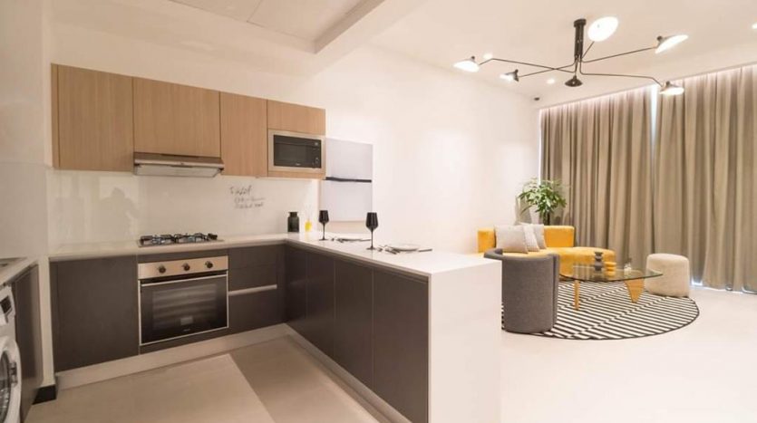 Une cuisine moderne avec des armoires grises, des accents en bois et des appareils électroménagers intégrés à l&#039;Aria Residence Dubai. Le salon adjacent comprend un fauteuil jaune, un canapé et un tapis rayé. Éclairage et rideaux lumineux