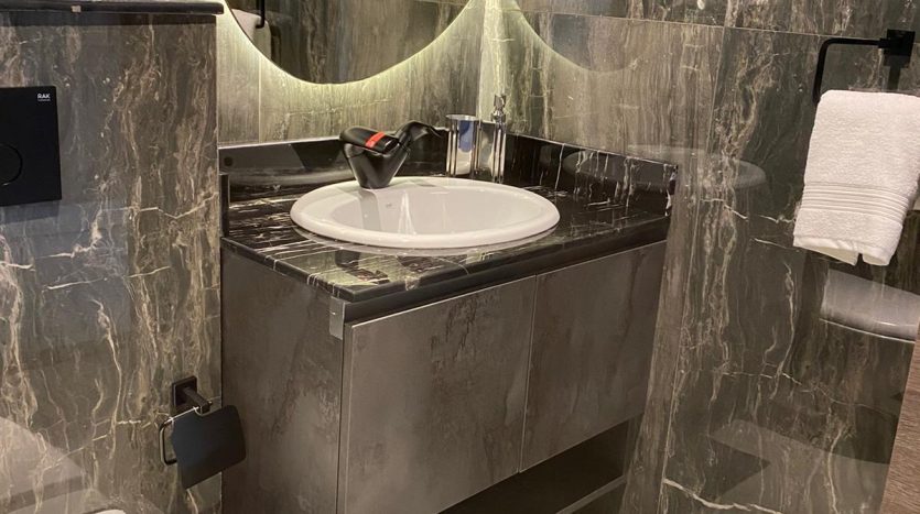 Une salle de bains moderne avec des murs et des surfaces en marbre sombre, avec un miroir circulaire au-dessus d&#039;un lavabo joya blanca, des toilettes à gauche et des serviettes soigneusement empilées.