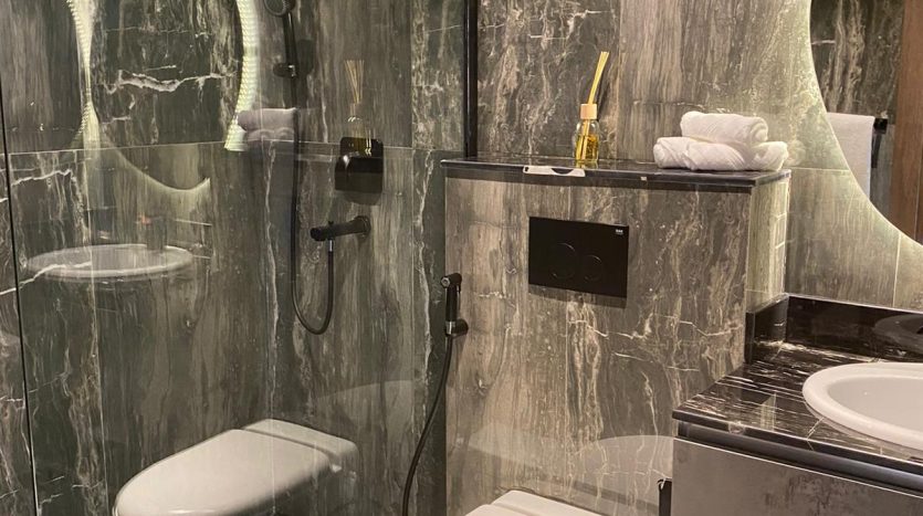 Une salle de bains moderne avec des murs et des sols en marbre foncé, avec un espace douche en verre transparent, des toilettes murales, un miroir sectionnel et des armoires grises Joya Blanca. Les serviettes et les articles de toilette sont