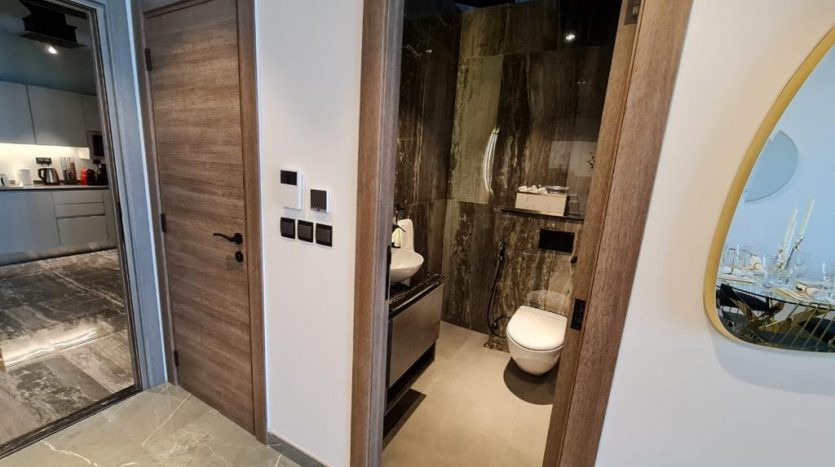 Intérieur de salle de bains moderne avec murs en marbre foncé, sol gris et portes marron partiellement ouvertes révélant des toilettes. Dans le reflet du miroir, un aperçu d&#039;une table à manger garnie de vaisselle est visible sur Joya Bl