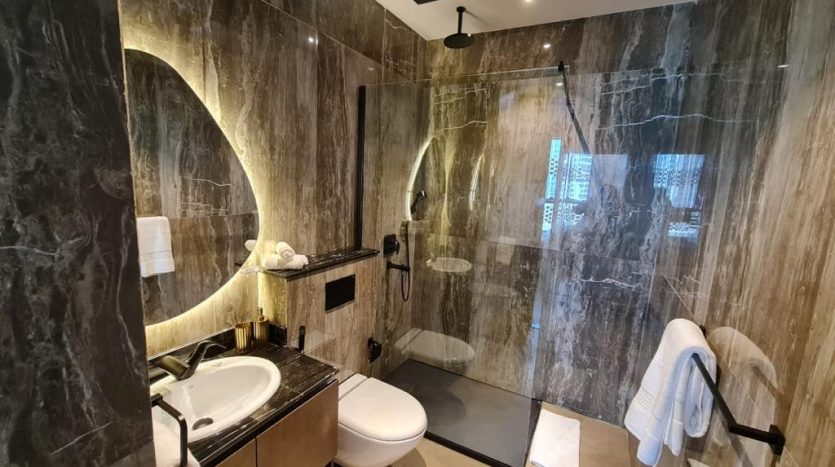 Une salle de bains moderne avec des murs et un sol en marbre foncé. Elle dispose d&#039;une douche à l&#039;italienne avec porte vitrée, d&#039;un lavabo Joya Blanca, d&#039;un miroir et de toilettes. Les serviettes sont bien rangées