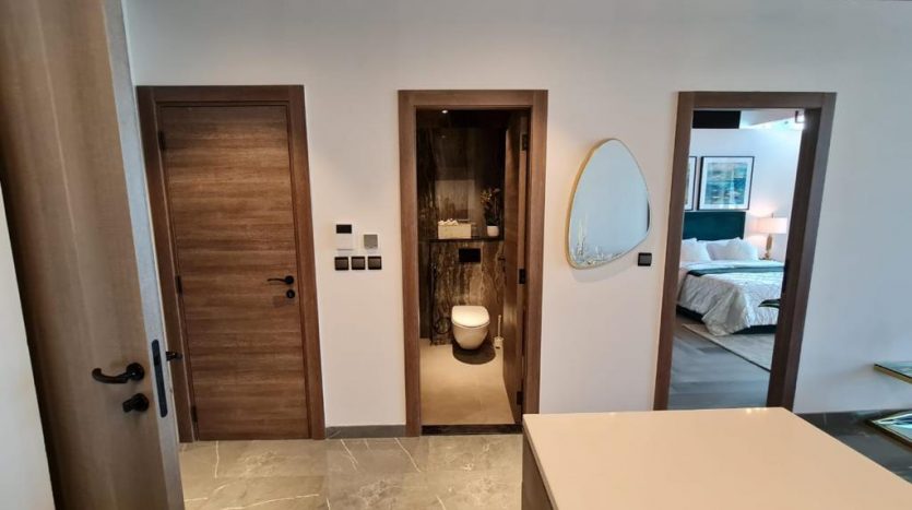 Un couloir intérieur moderne avec trois portes en bois, avec vue sur une salle de bain à gauche et une chambre joya blanca à droite. Un miroir décoratif et des œuvres d&#039;art sont exposés sur les murs.