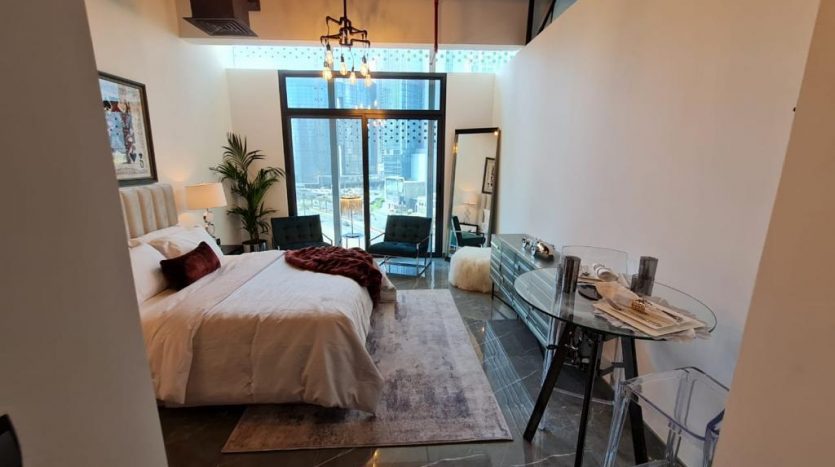 Un studio moderne avec un agencement ouvert comprenant un lit avec une literie blanche, des balcons en verre joya blanca avec vue sur la ville, un petit coin repas avec des chaises claires et un éclairage intérieur élégant.