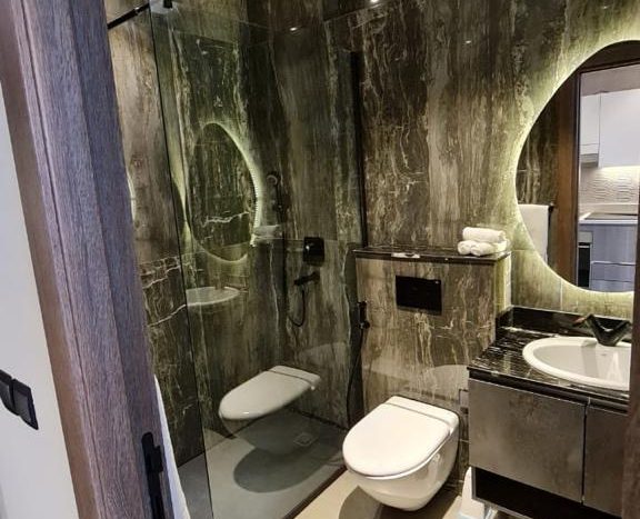 Une salle de bain moderne avec des murs en marbre foncé avec de grands miroirs, une douche à l&#039;italienne, des toilettes blanches et un lavabo joya blanca. Une serviette blanche est suspendue à côté du lavabo et le sol est
