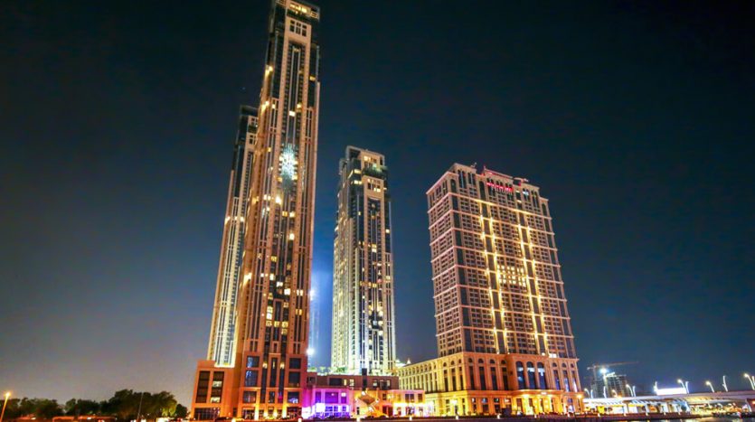 Un paysage urbain nocturne animé montrant trois grands bâtiments, dont la tour Amna, illuminés par des lumières colorées avec des reflets sur un plan d&#039;eau calme au premier plan dans la ville d&#039;Al Habtoor.