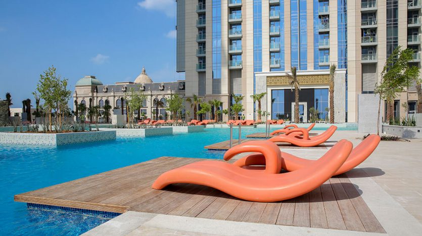 Espace piscine de l&#039;hôtel de luxe avec eau bleu clair et élégantes chaises longues orange sur la terrasse en bois, entourée de la tour Amna et de palmiers luxuriants sous un ciel bleu éclatant.