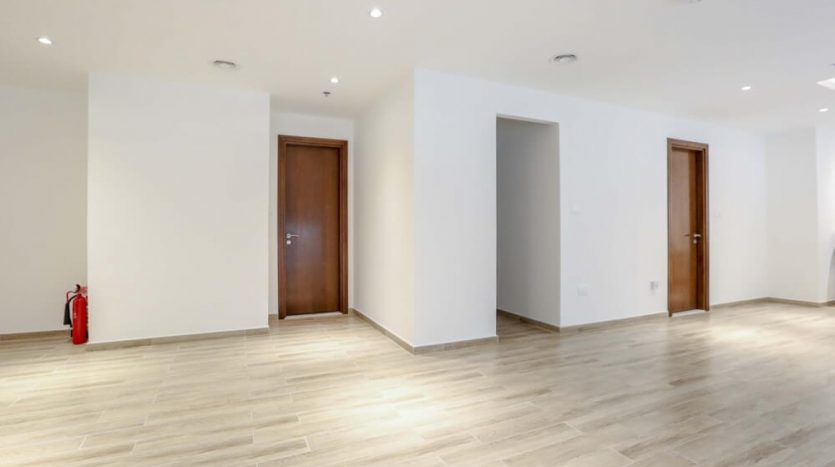 Une pièce spacieuse, moderne et vide dans la tour Amna à Al Habtoor City avec un parquet en bois clair, des murs blancs et de multiples portes en bois fermées. Un extincteur rouge se trouve à gauche