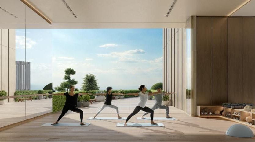 Trois personnes pratiquant le yoga dans une salle spacieuse et ensoleillée dotée de grandes fenêtres donnant sur un paysage extérieur serein à Dubaï. La chambre dispose de parquet et d&#039;une décoration minimaliste.