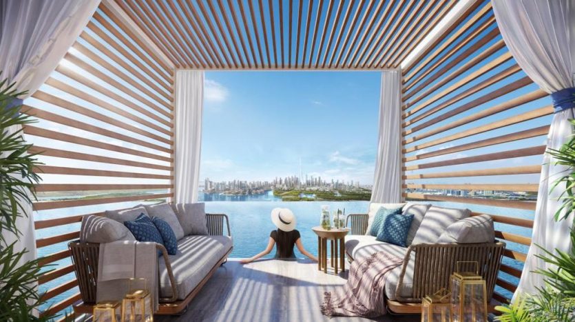 Une cabane luxueuse avec des rideaux blancs et des coussins rayés sur un canapé, surplombant les toits de la ville de Dubaï à travers un plan d&#039;eau. Une personne profite de la vue en portant un chapeau de soleil.
