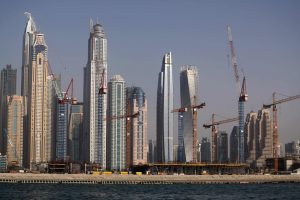 Horizonte de modernos rascacielos en construcción por un promotor inmobiliario en Dubai, con varias grúas, contra un cielo azul claro y visto desde el otro lado del agua.