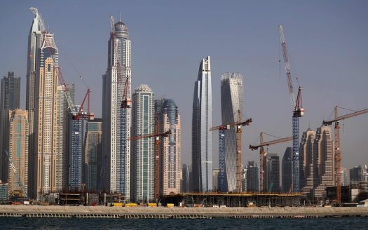 Horizon de gratte-ciel modernes en construction par un promoteur immobilier à Dubaï avec plusieurs grues, sur un ciel bleu clair et vu de l'autre côté de l'eau.