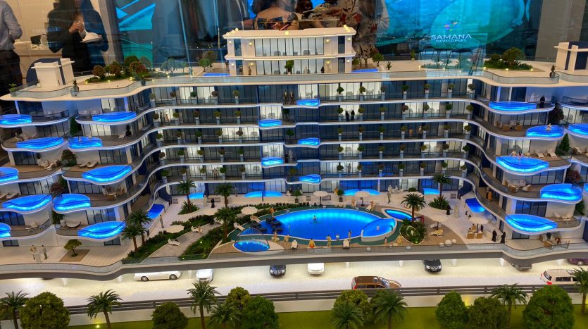 Un modèle détaillé de Samana Park Views, un complexe d&#039;appartements moderne avec une piscine centrale, des balcons éclairés en bleu, des palmiers environnants, des voitures miniatures sur la route et des personnes examinant le modèle