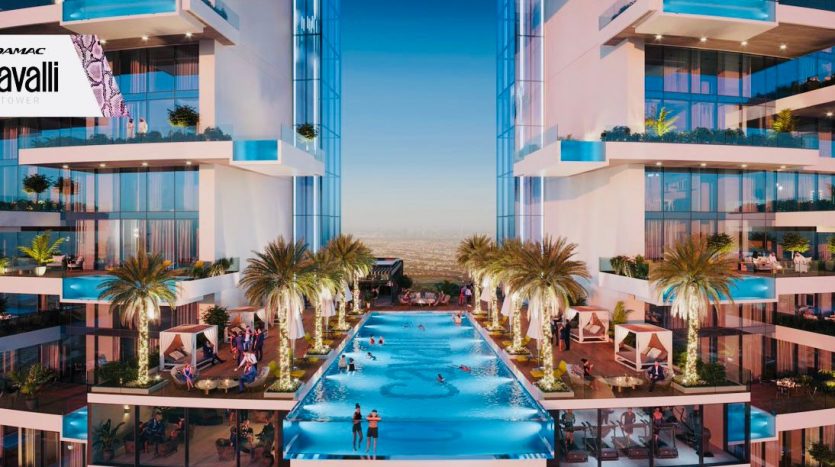 Une piscine luxueuse entre deux immeubles de grande hauteur, bordée de palmiers et de cabanes, sous un ciel dégagé. Les gens se détendent et nagent dans l’eau. Logos pour la tour Damac et Roberto Cavalli