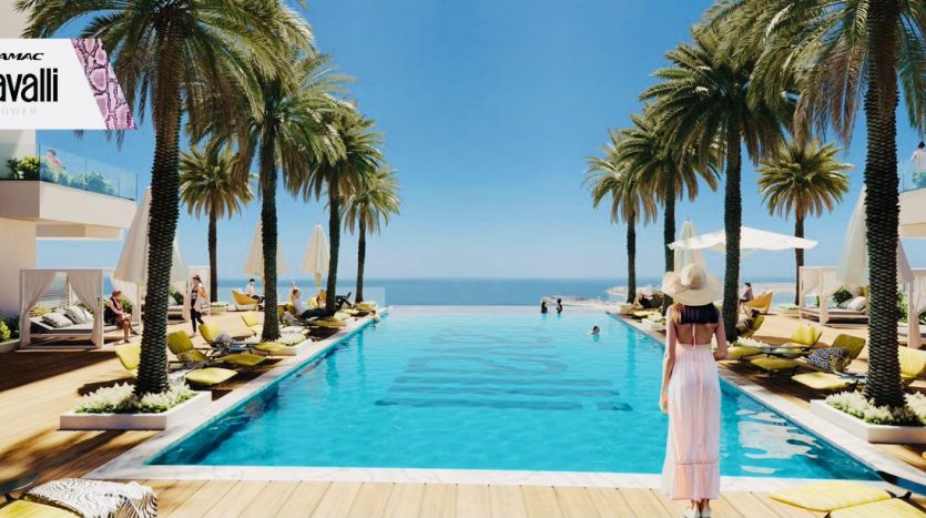 Une femme portant un chapeau et une robe fluide se tient au bord d&#039;une luxueuse piscine bordée de palmiers de la tour Roberto Cavalli à Dubaï, surplombant la mer. Des chaises longues et des parasols parsèment le