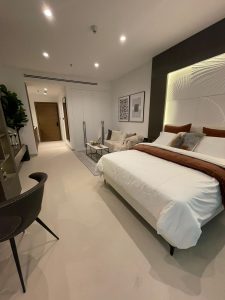Chambre moderne dans Prime Residency 3 avec un grand lit, une literie blanche et une tête de lit sombre. Les caractéristiques comprennent une table d'appoint, des œuvres d'art sur les murs, une chaise ronde et un éclairage élégant