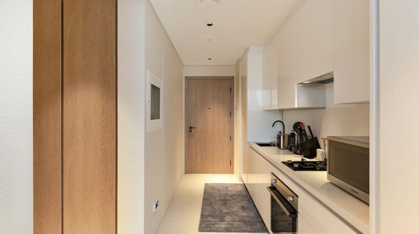 Une cuisine moderne et étroite avec des armoires en bois et une palette de couleurs beige, avec des appareils électroménagers intégrés et un couloir menant à une porte en bois clair à Marquise Square, Dubaï.