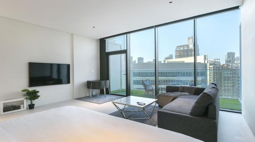 Salon d&#039;appartement moderne à Marquise Square, Dubaï avec de grandes fenêtres offrant une vue sur la ville, comprenant un canapé gris foncé, une table basse, une télévision montée et une décoration minimaliste.