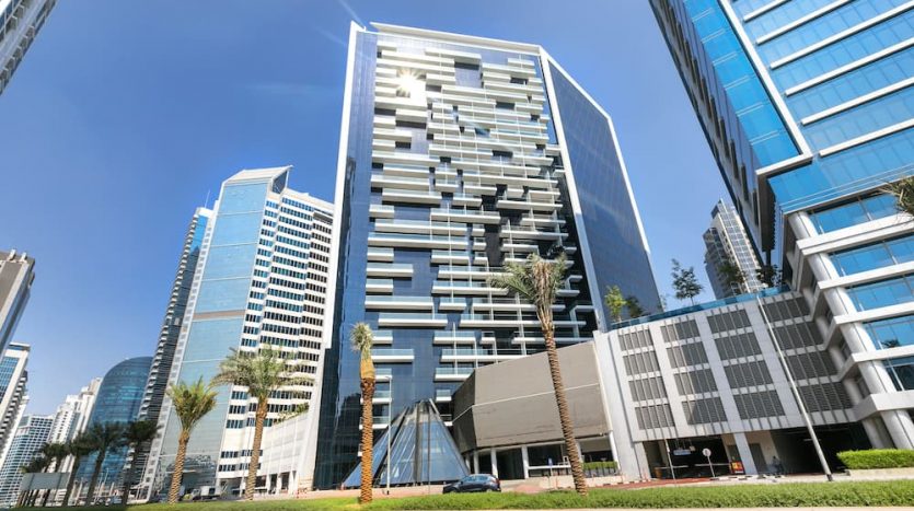 Paysage urbain moderne avec de grands gratte-ciel en verre et Marquise Square Dubai doté de balcons. Des palmiers luxuriants bordent la rue sous un ciel bleu clair.