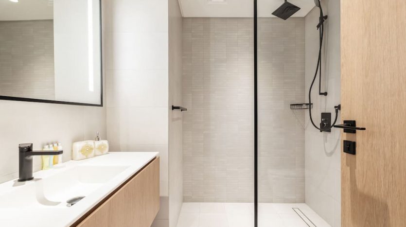 Salle de bains moderne dotée d&#039;un comptoir blanc élégant avec un lavabo, un miroir éclairé et une douche à l&#039;italienne spacieuse avec une pomme de douche noire et une porte vitrée sur la place Marquise, à Dubaï.