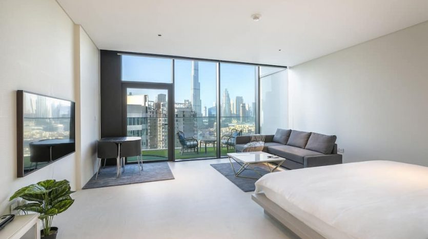 Chambre moderne à Marquise Square, Dubaï avec un grand lit, un canapé et une télévision, dotée de grandes fenêtres qui mettent en valeur les toits de la ville. La chambre présente une décoration minimaliste et un balcon.