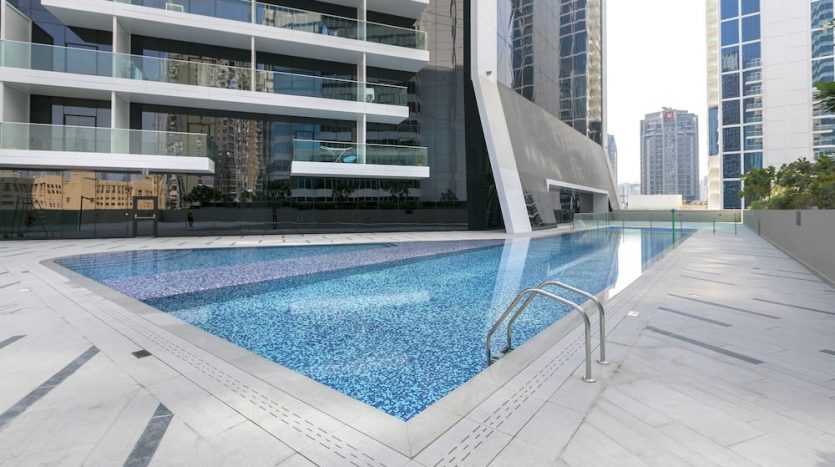 Une piscine extérieure avec des carreaux de mosaïque bleue, située sur Marquise Square Dubaï, entourée d&#039;immeubles modernes de grande hauteur, dotée d&#039;une terrasse propre et ouverte dans un environnement urbain.