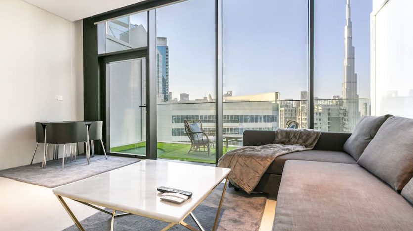 Salon moderne avec de grandes fenêtres en verre offrant une vue spectaculaire sur les toits de Marquise Square Dubaï, y compris un gratte-ciel proéminent. La pièce comprend un canapé sectionnel, une table basse blanche et un décor minimaliste.